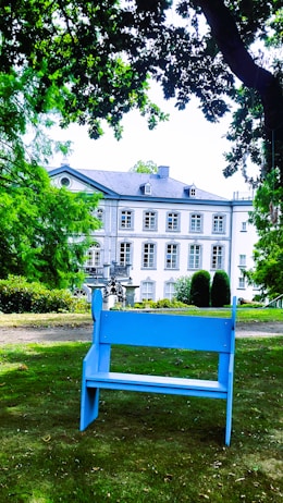 A serene garden setting featuring a blue bench prominently placed in the foreground. The backdrop consists of a grand, elegant building with multiple windows and a light-colored facade. Lush greenery, including trees and bushes, surrounds the area, contributing to a peaceful and inviting atmosphere.