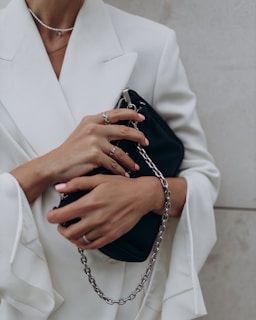 A person wearing a white blazer holds a black clutch purse with a silver chain. Their fingers are adorned with rings, and their nails are manicured with a pale pink polish.