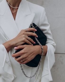 A person wearing a white blazer holds a black clutch purse with a silver chain. Their fingers are adorned with rings, and their nails are manicured with a pale pink polish.