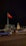 A large red flag is prominently displayed on a flagpole, with a modern building in the background. To the right of the flag is a statue of a person on horseback, illuminated by lights. The scene is set at night, with moving vehicles on a street in the foreground and low lighting creating a calm atmosphere.