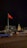A large red flag is prominently displayed on a flagpole, with a modern building in the background. To the right of the flag is a statue of a person on horseback, illuminated by lights. The scene is set at night, with moving vehicles on a street in the foreground and low lighting creating a calm atmosphere.