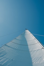 A large, white sail is captured from below against a clear blue sky. The angle provides a dynamic view of the sail’s curves and rigging lines, creating a sense of openness and movement.