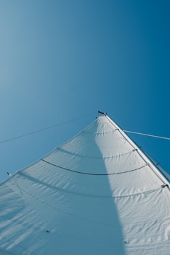 A large, white sail is captured from below against a clear blue sky. The angle provides a dynamic view of the sail’s curves and rigging lines, creating a sense of openness and movement.