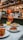 A well-set table with several appetizing dishes, including pasta with sauce, a layered lasagna, a beetroot salad, and other colorful meals. At the center, there's a tall glass of Aperol Spritz with ice, emanating a vibrant orange hue. In the background, a bar area with shelves filled with bottles and hanging wine glasses creates a lively ambiance.