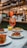 A well-set table with several appetizing dishes, including pasta with sauce, a layered lasagna, a beetroot salad, and other colorful meals. At the center, there's a tall glass of Aperol Spritz with ice, emanating a vibrant orange hue. In the background, a bar area with shelves filled with bottles and hanging wine glasses creates a lively ambiance.