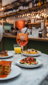 A well-set table with several appetizing dishes, including pasta with sauce, a layered lasagna, a beetroot salad, and other colorful meals. At the center, there's a tall glass of Aperol Spritz with ice, emanating a vibrant orange hue. In the background, a bar area with shelves filled with bottles and hanging wine glasses creates a lively ambiance.