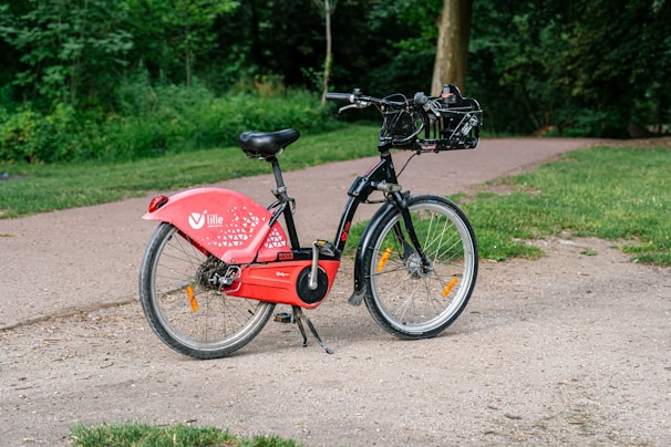 A red and black bicycle is parked on a gravel path near a grassy area. The bike has a labeled panel and includes a basket on the handlebars. Surrounding the path are trees and greenery, creating a natural setting.