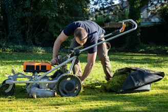 A person is kneeling on a grassy lawn, operating a lawn mower. The person is wearing a dark shirt and khaki pants. The lawn mower is metallic with orange accents and has a grass collection bag attached to it. In the background, there are trees and shrubs providing a lush, green backdrop.
