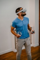A man wearing a virtual reality headset stands indoors on a wooden floor, holding two VR controllers, looking slightly upwards. He wears a blue shirt and beige pants.