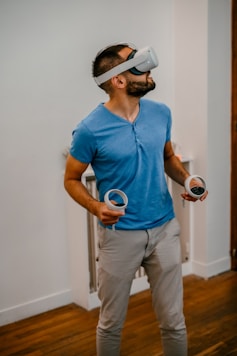 A man wearing a virtual reality headset stands indoors on a wooden floor, holding two VR controllers, looking slightly upwards. He wears a blue shirt and beige pants.