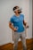 A man wearing a virtual reality headset stands indoors on a wooden floor, holding two VR controllers, looking slightly upwards. He wears a blue shirt and beige pants.