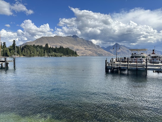 A serene lakeside scene features calm water with a floating bar docked at a pier. Majestic mountains rise in the background beneath a dramatic sky filled with fluffy clouds. Trees line the shore, adding a touch of greenery.