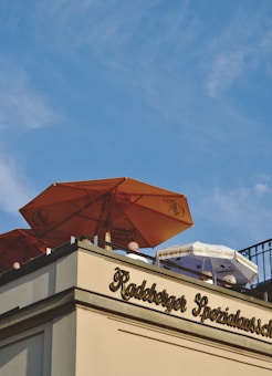A rooftop terrace features large orange and white umbrellas with branding, partially covering people seated below. The building's facade displays ornate lettering with the name 'Radeberger'. Above, a bright blue sky dominates the scene.