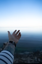 A tattooed arm extended outward against a serene landscape, with a gradient blue sky meeting a distant horizon over a body of water or possibly a misty valley. The foreground shows a portion of a striped sleeve.