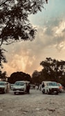 A parking area with several white vans and people walking around. Trees are visible in the background under a sky filled with dramatic clouds and a hint of sunset colors.