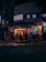 A street scene at night featuring a small outdoor restaurant or food stall with people seated at tables. The establishment is adorned with Korean signage and has a cozy, illuminated interior that contrasts with the darker street surroundings.