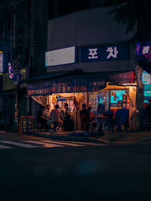 A street scene at night featuring a small outdoor restaurant or food stall with people seated at tables. The establishment is adorned with Korean signage and has a cozy, illuminated interior that contrasts with the darker street surroundings.
