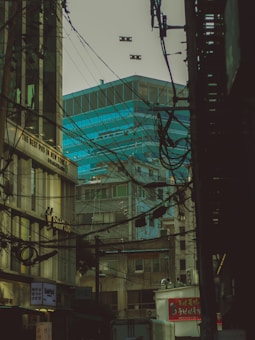 The image depicts an urban alleyway or street dense with overhead wires and utility cables. In the background, modern glass buildings with a blue tint contrast with older, more worn buildings in the foreground. Various signs, some possibly in local languages, are present and a vehicle is visible at the bottom of the frame. The scene is enveloped in dim lighting, casting an overall shadowy and cluttered atmosphere.