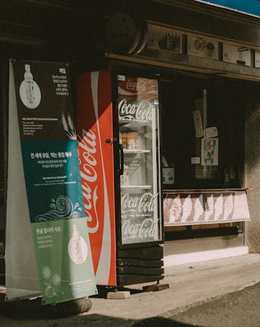 A Coca-Cola branded refrigerated display case stands outside a small storefront. A promotional banner with Korean text and images hangs beside it. Various items are visible inside the refrigerator. The store has a rustic appearance with signage above the entrance.