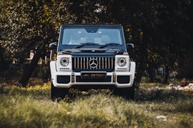A white and black luxury SUV with a distinctive grille is parked on grass, surrounded by trees, creating a natural backdrop. The vehicle's brand logo is prominently displayed on the front.