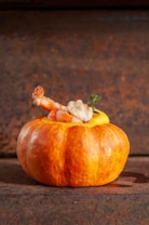 A small pumpkin serves as a bowl, filled with a creamy, yellow soup garnished with shrimp and a sprig of thyme. The background is a textured, rustic brown surface.