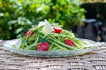 A plate of fresh zucchini noodles is topped with vibrant cherry tomato halves, shavings of parmesan cheese, and small basil leaves. The dish is presented on a clear glass plate with a woven rattan texture surface underneath. The background is a blurred garden scene with lush greenery.