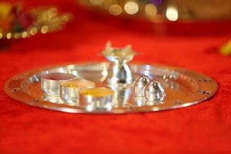 A silver tray with intricate designs holds several small metal vessels containing colorful powders. The tray is placed on a vibrant red cloth that creates a warm and festive atmosphere.