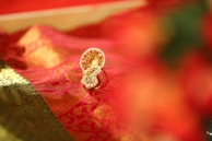 A detailed golden and red embellished fabric is draped elegantly, featuring intricate patterns and textures. Nestled on the fabric is an ornate gold ornament with small embedded pearls and red stones, exuding luxury and cultural richness.