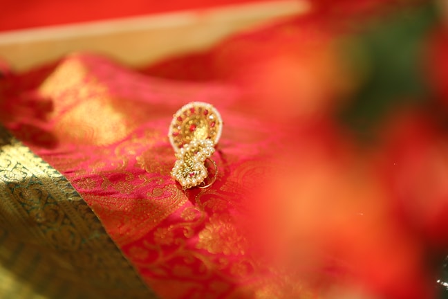 A detailed golden and red embellished fabric is draped elegantly, featuring intricate patterns and textures. Nestled on the fabric is an ornate gold ornament with small embedded pearls and red stones, exuding luxury and cultural richness.