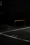 A dimly lit parking area featuring a yellow metal barrier positioned in the center of a parking space marked with the number 29. The ground is covered with asphalt and the background includes horizontal slatted structures partially visible in low light.