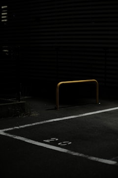A dimly lit parking area featuring a yellow metal barrier positioned in the center of a parking space marked with the number 29. The ground is covered with asphalt and the background includes horizontal slatted structures partially visible in low light.