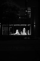 Four mannequins wearing elegant wedding dresses are displayed in the window of a store, illuminated by soft lighting in an otherwise dark environment. The view captures the detailed design of the dresses against the glass, with reflections creating an atmospheric contrast.