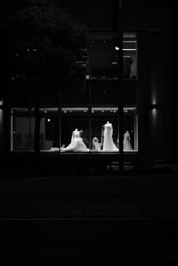 Four mannequins wearing elegant wedding dresses are displayed in the window of a store, illuminated by soft lighting in an otherwise dark environment. The view captures the detailed design of the dresses against the glass, with reflections creating an atmospheric contrast.