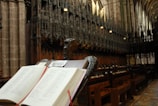 An open Bible rests on a wooden lectern with an ornate carved design. The setting is a church interior with large stone columns and intricate wooden choir stalls lining the sides. The space is dimly lit, creating a serene atmosphere with a sign that reads 'PLEASE DO NOT TOUCH THE BIBLE' visible on the lectern.
