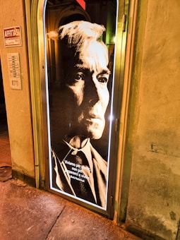 A portrait with a vintage aesthetic featuring a serious-looking individual in formal attire. The monochrome style with high contrast emphasizes the facial features and gives a dramatic tone. An Italian quote by Boris Pasternak is displayed at the bottom. The image is mounted on a door illuminated by a halo of light.