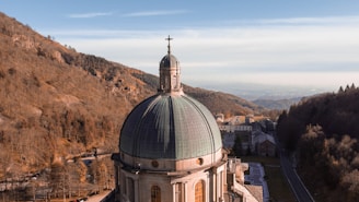 A large church dome with a cross stands prominently against a backdrop of forested hills. The dome features a greenish patina and intricate architectural details. The landscape extends into the distance with a few buildings visible and a winding road entering a wooded area.