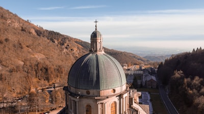 A large church dome with a cross stands prominently against a backdrop of forested hills. The dome features a greenish patina and intricate architectural details. The landscape extends into the distance with a few buildings visible and a winding road entering a wooded area.
