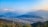 A panoramic view of a valley with a city nestled between lush green hills and majestic snow-capped mountains in the distance. The sky is clear and blue, with a warm golden light casting over the scene.