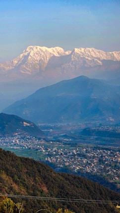 A breathtaking landscape featuring snow-capped mountains in the distance, with lush green hills and a sprawling village in the foreground. The image captures the serene beauty of nature and human habitation coexisting harmoniously.