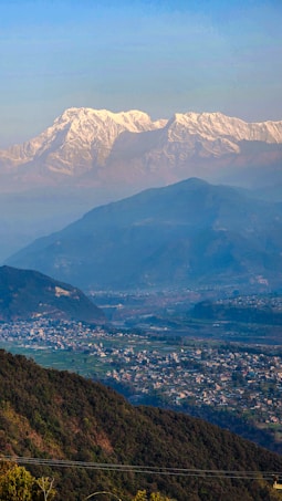 A breathtaking landscape featuring snow-capped mountains in the distance, with lush green hills and a sprawling village in the foreground. The image captures the serene beauty of nature and human habitation coexisting harmoniously.