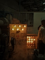 An industrial setting with various machinery. There is a control panel with several glowing bulbs and meters. A rack with multiple glowing lights is placed next to it. A person is partially visible on the right side of the image. A smoky or foggy atmosphere adds a dim ambiance to the scene.