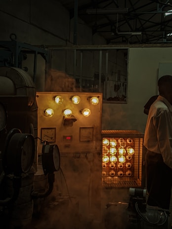 An industrial setting with various machinery. There is a control panel with several glowing bulbs and meters. A rack with multiple glowing lights is placed next to it. A person is partially visible on the right side of the image. A smoky or foggy atmosphere adds a dim ambiance to the scene.
