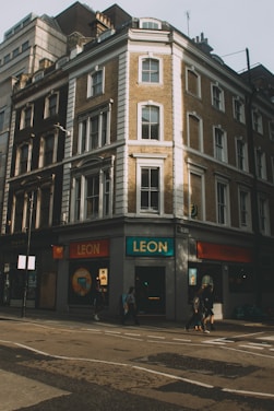A multi-story corner building with classic architectural features, such as large windows and decorative elements, displaying a restaurant sign with the name 'Leon' at the ground level. Several pedestrians are walking along the street, and the overall scene appears to be taken during daytime, with shadows indicating the angle of the sun.