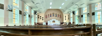 A spacious church interior featuring long wooden pews under a high, bright ceiling. Colorful stained glass windows line the walls, and religious artwork is displayed above the altar. Ceiling fans and speakers are mounted along the walls.