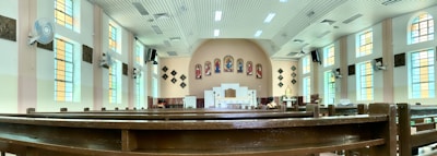 A spacious church interior featuring long wooden pews under a high, bright ceiling. Colorful stained glass windows line the walls, and religious artwork is displayed above the altar. Ceiling fans and speakers are mounted along the walls.