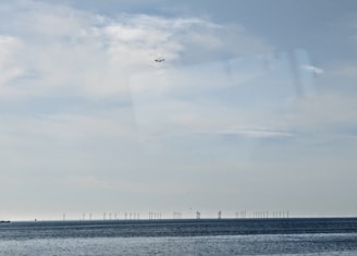 A serene seascape featuring a calm body of water stretching towards the horizon. Wind turbines are visible in the distance beneath a sky that is predominantly overcast with patches of blue. A small airplane is flying high in the sky.