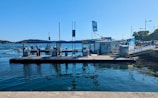 A waterfront scene featuring a fuel dock on a clear day. The dock is equipped with several fuel pumps and is positioned in a harbor area. A small building with colorful flags on the roof is visible, and beyond the dock, calm water extends towards a distant treeline. Boats can be seen in the background, along with clear blue skies overhead.
