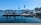 A waterfront scene featuring a fuel dock on a clear day. The dock is equipped with several fuel pumps and is positioned in a harbor area. A small building with colorful flags on the roof is visible, and beyond the dock, calm water extends towards a distant treeline. Boats can be seen in the background, along with clear blue skies overhead.