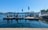 A waterfront scene featuring a fuel dock on a clear day. The dock is equipped with several fuel pumps and is positioned in a harbor area. A small building with colorful flags on the roof is visible, and beyond the dock, calm water extends towards a distant treeline. Boats can be seen in the background, along with clear blue skies overhead.