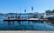 A waterfront scene featuring a fuel dock on a clear day. The dock is equipped with several fuel pumps and is positioned in a harbor area. A small building with colorful flags on the roof is visible, and beyond the dock, calm water extends towards a distant treeline. Boats can be seen in the background, along with clear blue skies overhead.